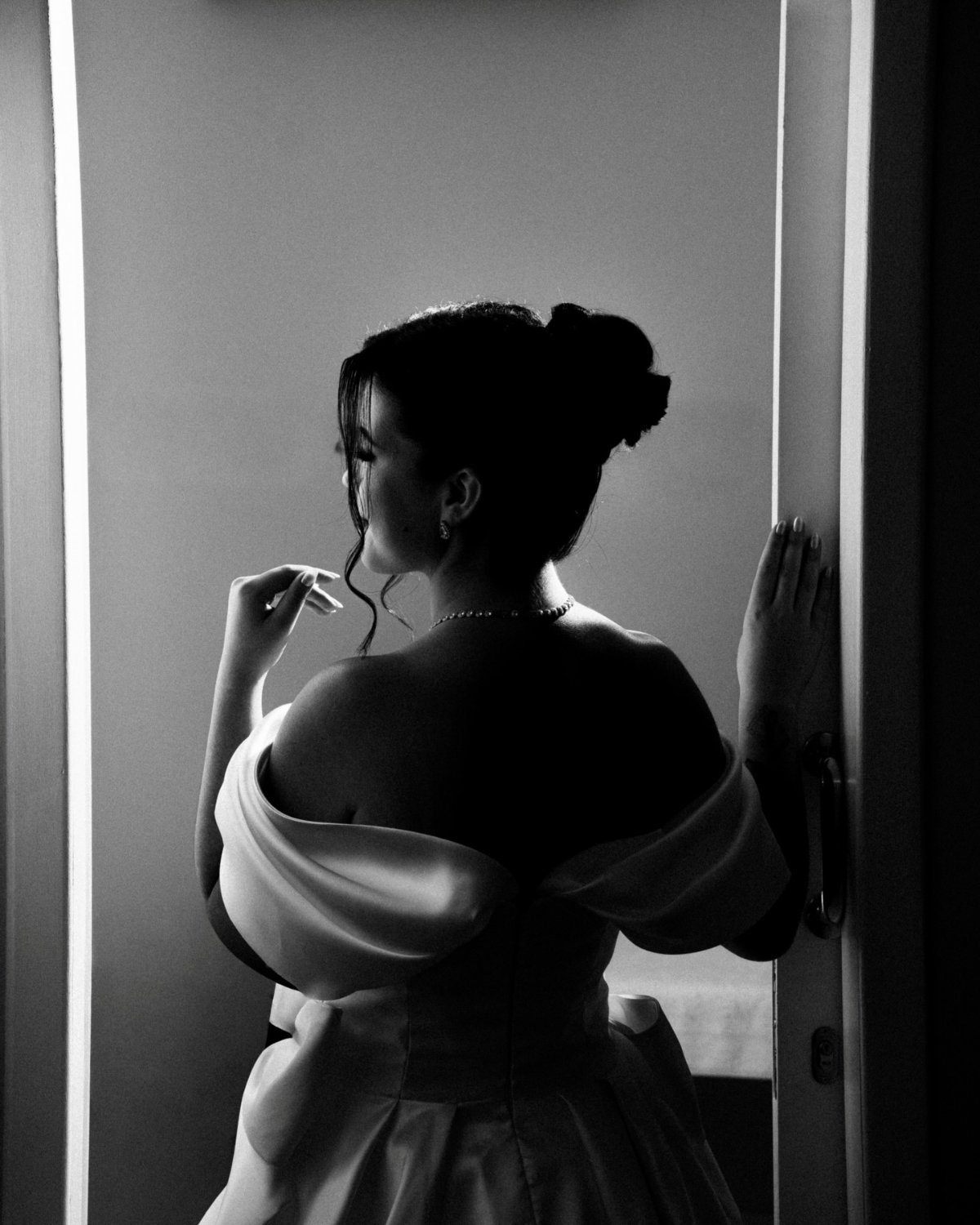 Noiva de costas junto a uma porta, em contraluz, em preto e branco.